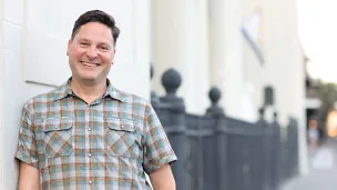 Man in plaid shirt smiling against building columns.