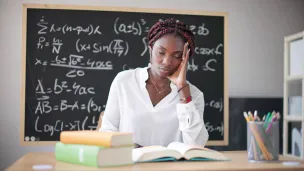 Woman at desk with books and math formulas on blackboard.