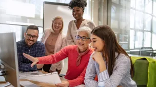 A diverse group of five smiling colleagues, three women and two men, are gathered around a computer in a modern office. A seated woman points at the screen, engaging the group in a lively discussion.