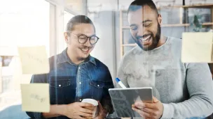 Two men laughing while looking at a tablet, with sticky notes on a glass wall.