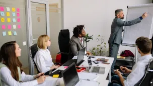 Man in suit presents on whiteboard to diverse group in meeting room.