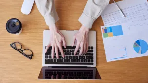 Hands typing on a laptop next to a coffee cup, glasses, and charts.