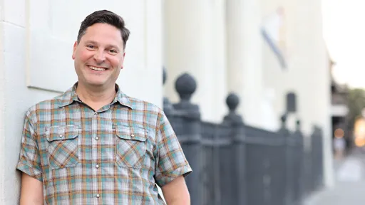 Man in plaid shirt smiling against building columns.