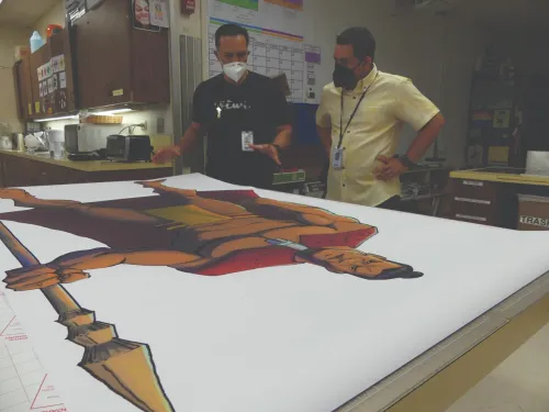 Two men in masks look at a print of a Hawai'ian warrior.