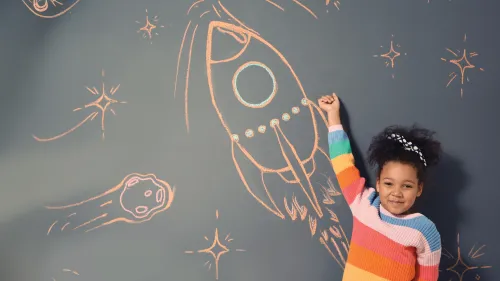 Child drawing a rocket with chalk on a dark wall.