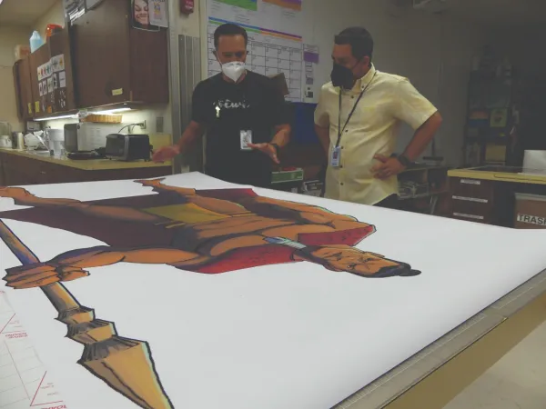 Two men in masks look at a print of a Hawai'ian warrior.